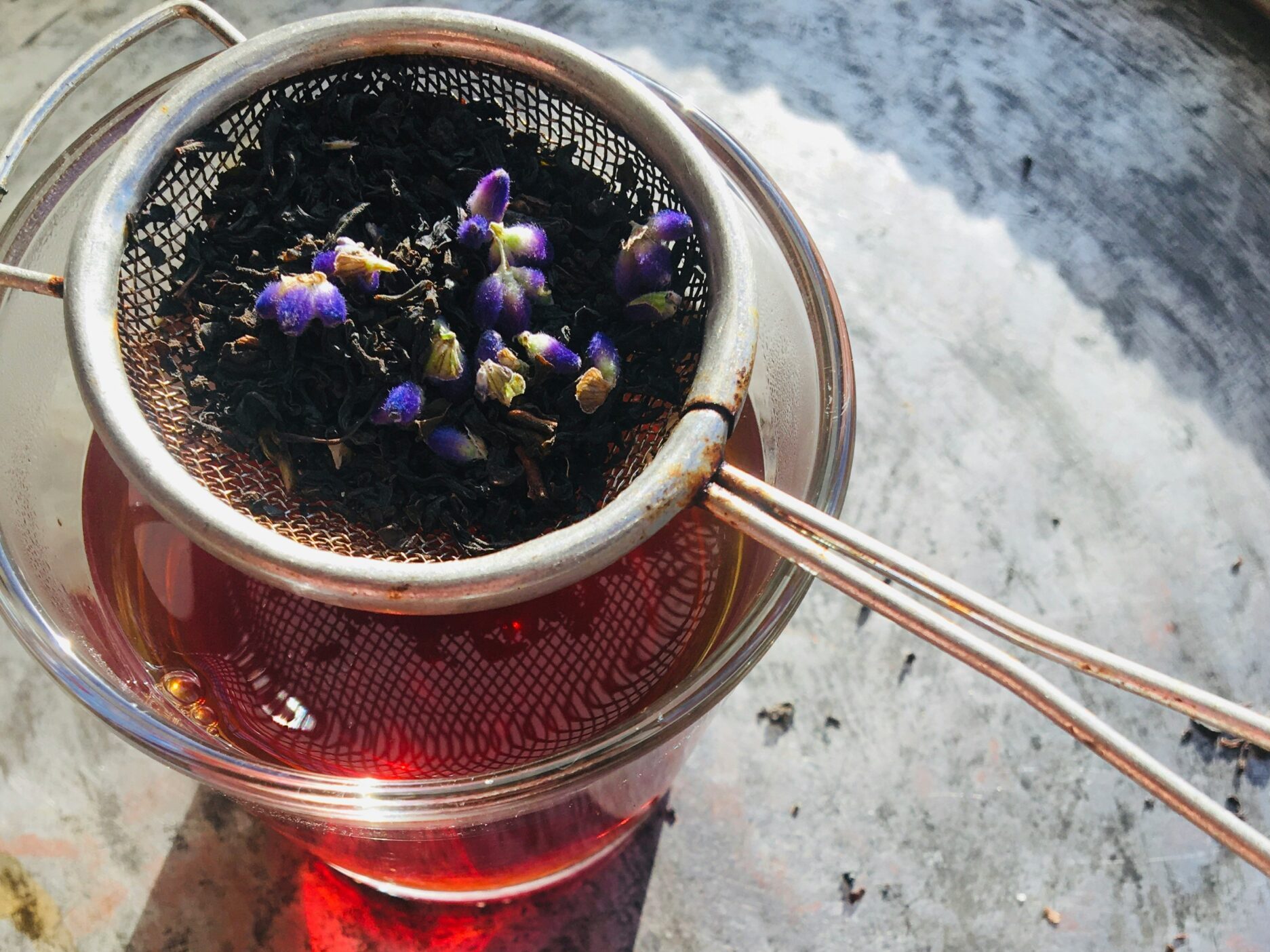 A close-up of a tea infuser containing black tea and lavender buds, sitting atop a glass teacup filled with deep red tea. Fresh lavender sprigs are placed beside the cup on a textured surface, highlighting the calming ambiance of herbal tea preparation.
